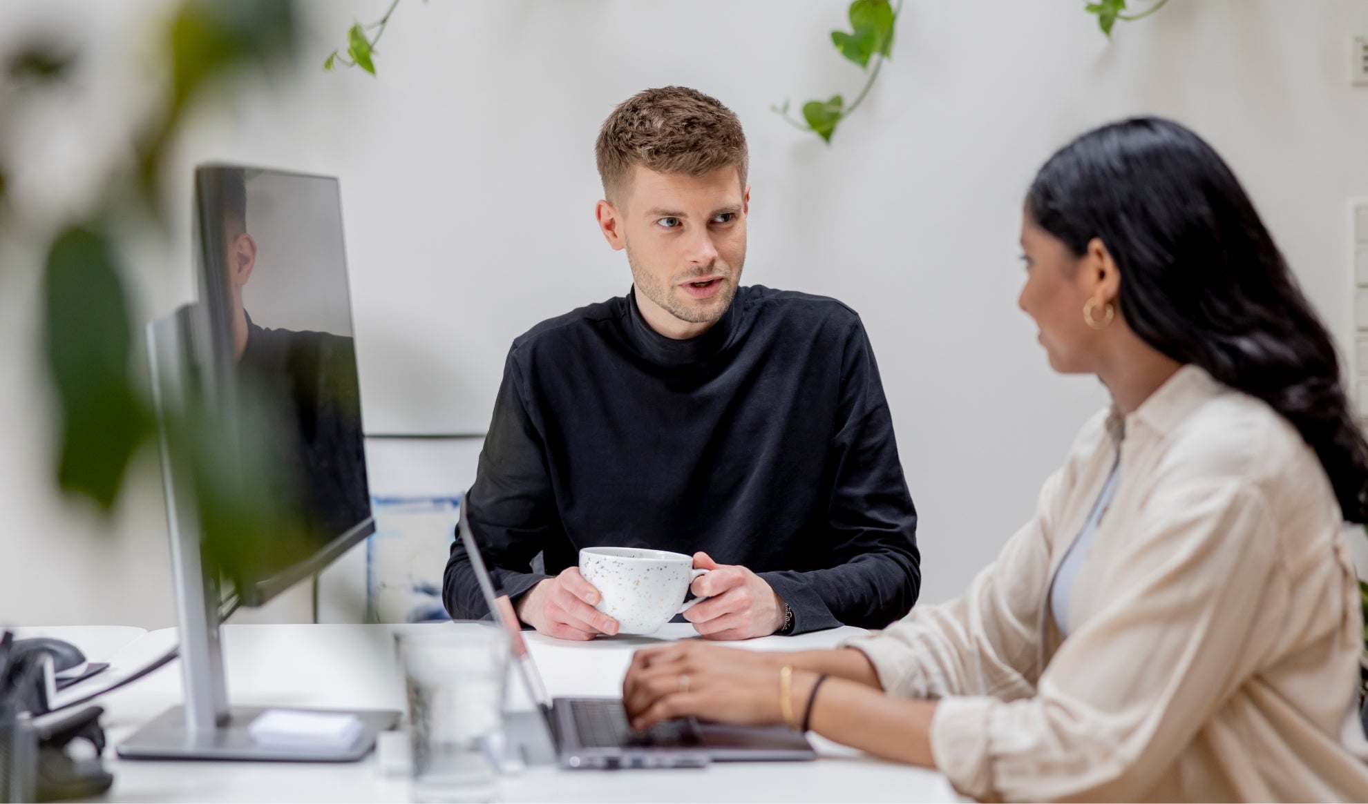 Zwei Kollegen am Schreibtisch im hellen Büro unterhalten sich eine tippt am Laptop andere hält Kaffeetasse