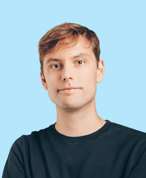 junger Mann mit kurzen hellbraunen Haaren und neutralem Ausdruck in schwarzem T-Shirt vor hellblauem Hintergrund