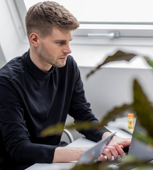 junger Mann sitzt konzentriert vor Laptop in hellem Büro mit Pflanzen im Vordergrund