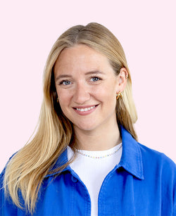 junge blonde Frau lächelt vor rosafarbenem Hintergrund trägt blaue Jacke goldene Creolen
