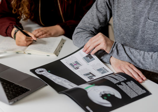 Zwei Personen lesen am Schreibtisch technische Broschüre und notieren handgeschriebene Notizen im Notizbuch neben Laptop