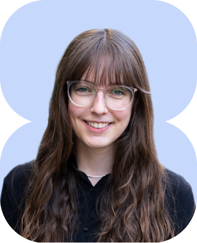 lächelnde junge Frau mit langen braunen Haaren und geradem Pony, filigrane Brille, schwarzes Hemd, pastellblauer Hintergrund