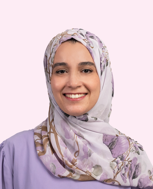 junge Frau mit gemustertem Hijab und lila Bluse lächelt vor pastellrosa Hintergrund