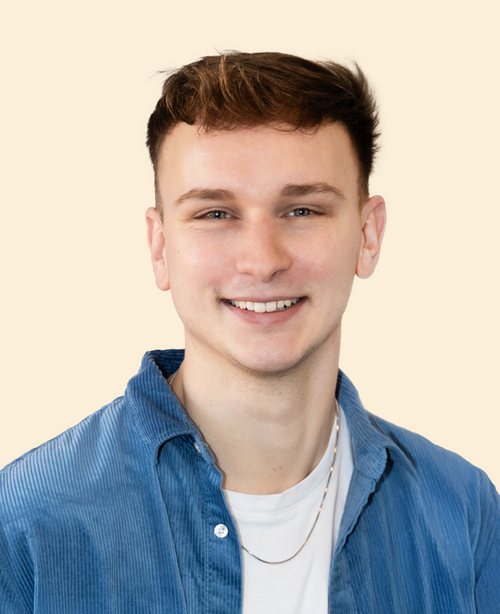 Junger Mann mit kurzen braunen Haaren und blauem Cordhemd lächelt vor beigem Hintergrund