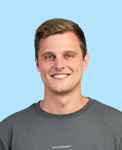 lächelnder Mann mit kurzem braunem Haar grauem Pullover mit Schriftzug Daily Aesthetikz vor pastellblauem Hintergrund
