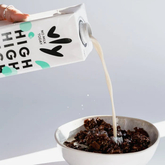 Hand gießt pflanzenmilch aus Karton über knuspriges Schokoladenmüsli in weißer Schale