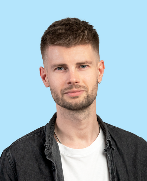 junger Mann mit kurzem braunen Haar, leichtem Bart, silberner Kette und schwarzer Jeansjacke vor hellblauem Hintergrund