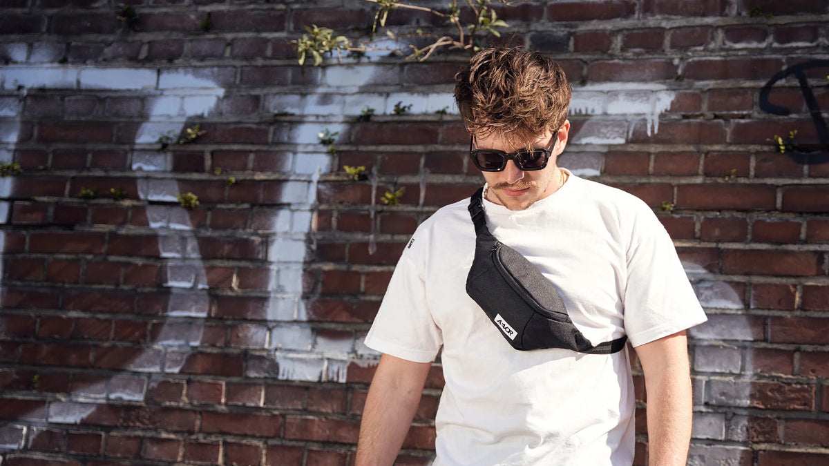 Junger Mann mit schwarzer Bauchtasche und dunkler Sonnenbrille vor graffitiverzierter Backsteinmauer