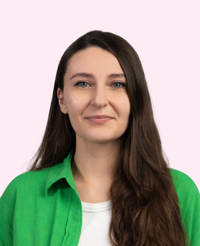 Frau mit langen braunen Haaren, neutral lächelnd, grünem Hemd vor pastellrosa Hintergrund