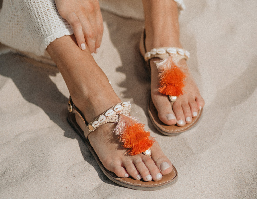 Füße in Bastsandalen mit Muscheln und orangefarbenen Quasten auf Sand
