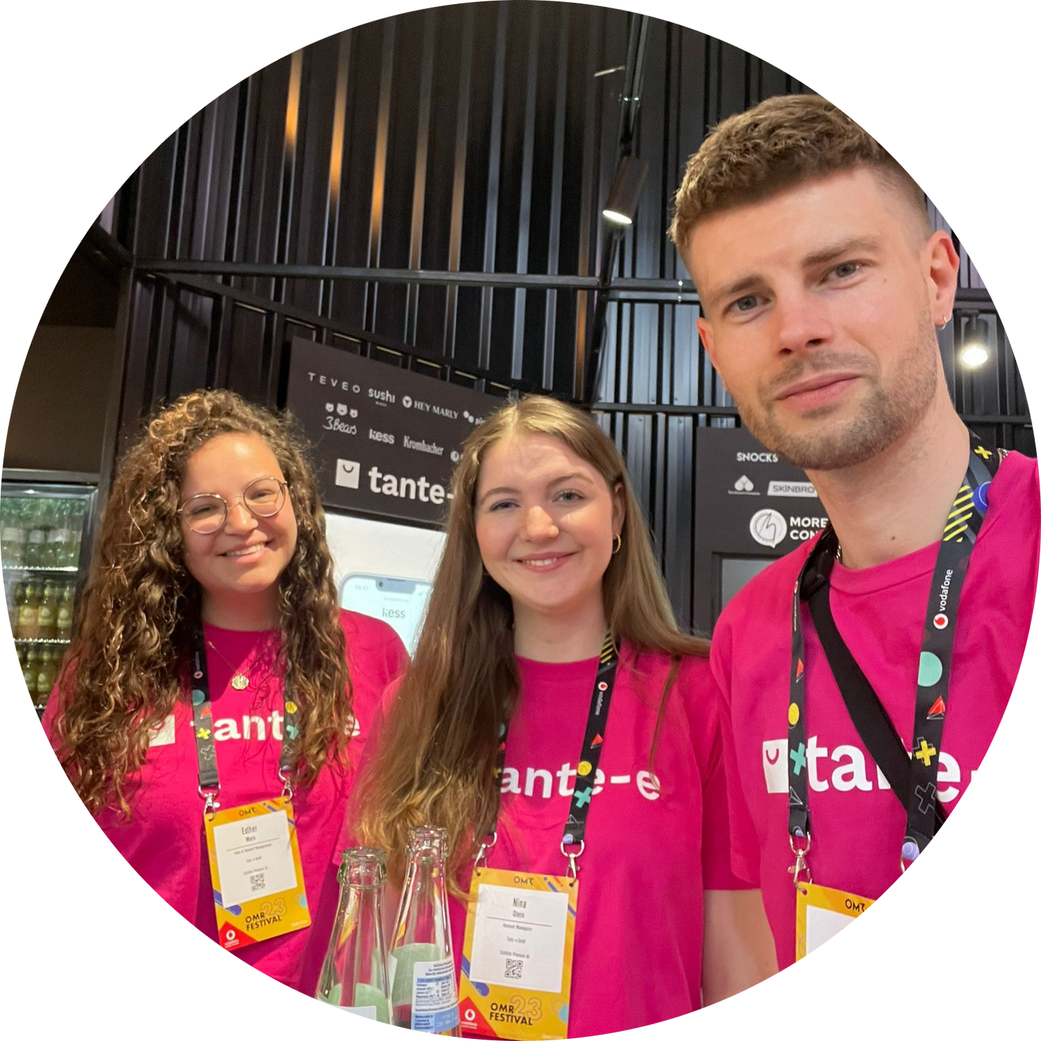 Drei junge Tante-e-Mitarbeitende in pinken Shirts lächeln vor Messestand mit Getränkeflaschen und Namensschildern