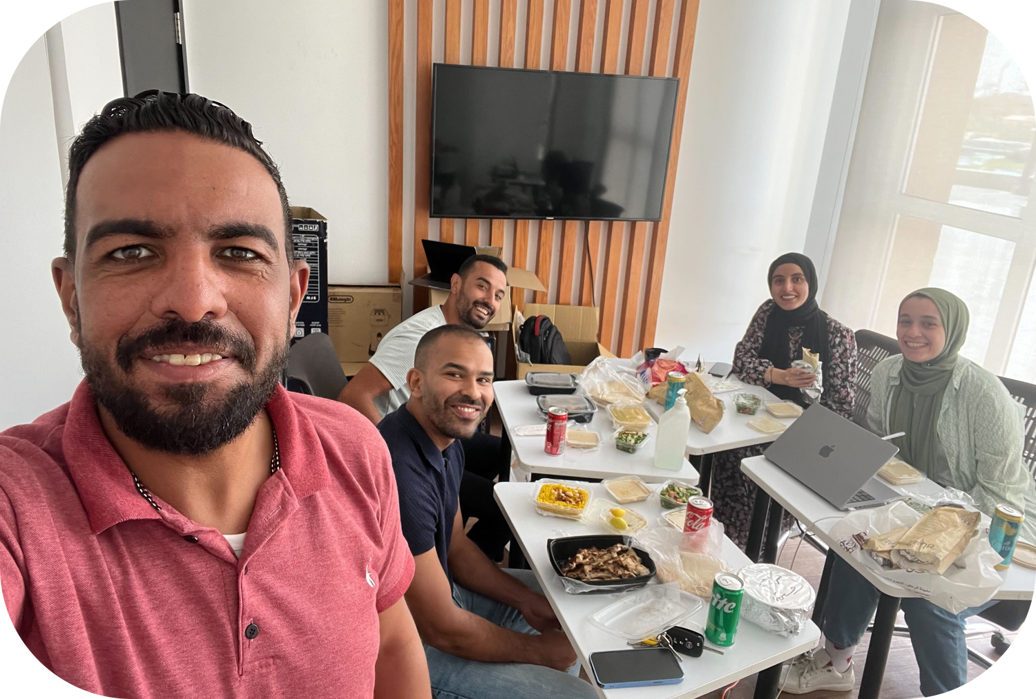 Fünf Personen sitzen am Tisch mit Lunchpaketen und Getränken vor Fernsehbildschirm in modernem Büro