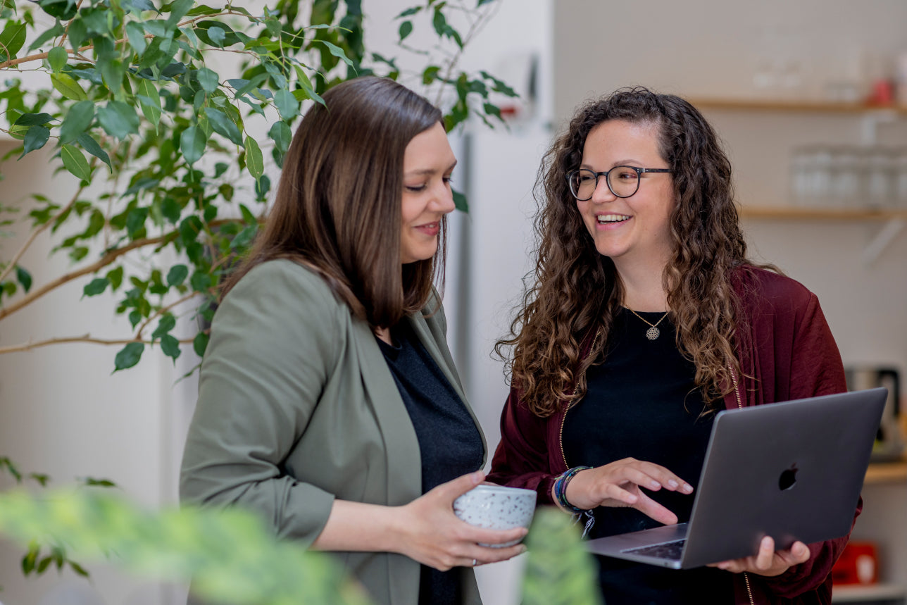 Zwei lächelnde Frauen mit Laptop und Kaffeetasse in heller Büroatmosphäre mit Zimmerpflanzen im Hintergrund