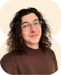 Junge Person mit lockigem dunklem Haar und klarem Brillengestell trägt braunen Strickpullover lächelnd vor hellem Hintergrund