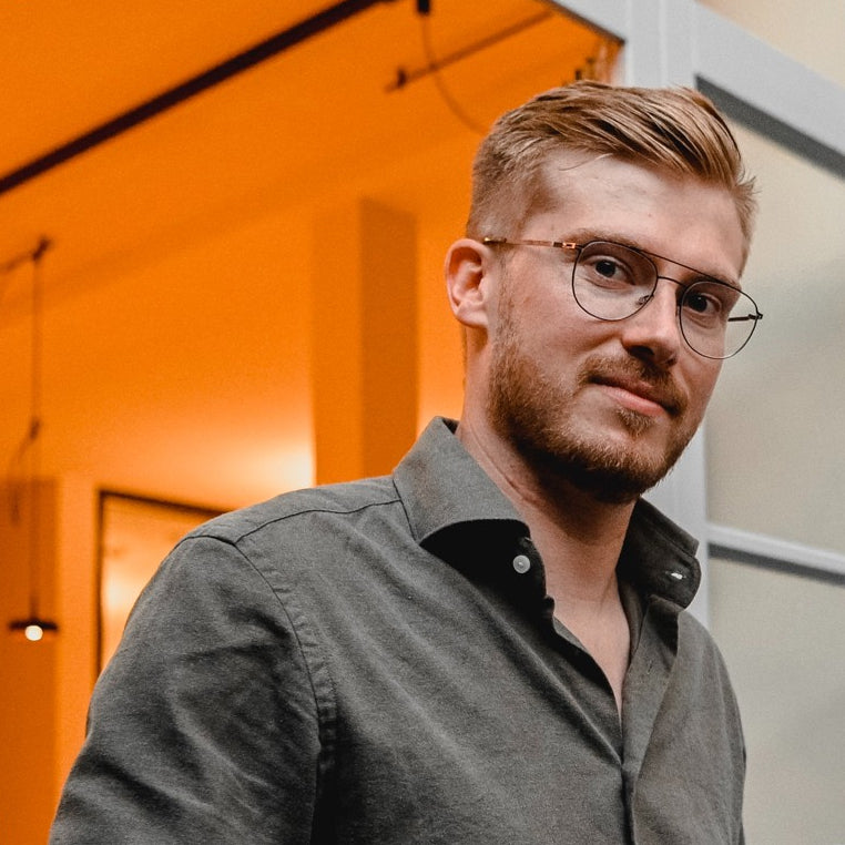 junger Mann mit Brille und Bart in grauem Hemd im orange beleuchteten Raum vor Glaswand