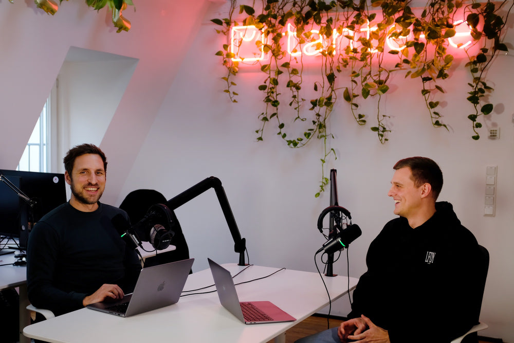 Zwei lächelnde Männer sitzen an einem Tisch mit Laptops und Mikrofonen in modernem Büro mit hängenden Pflanzen und Neonlicht
