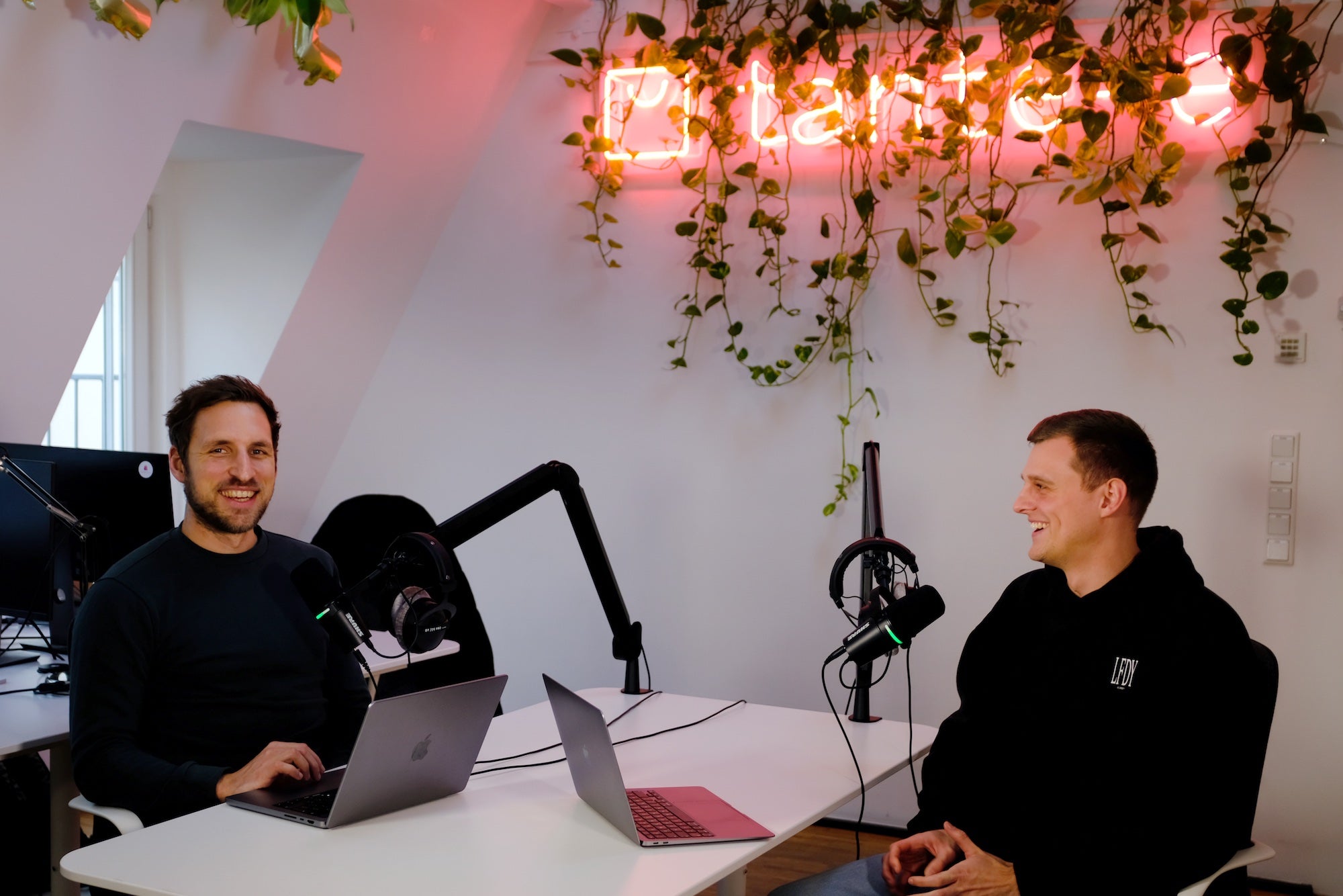 Zwei lächelnde Männer sitzen an einem Tisch mit Laptops und Mikrofonen in modernem Büro mit hängenden Pflanzen und Neonlicht