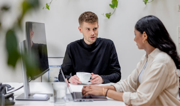 Zwei Kollegen am Schreibtisch im hellen Büro unterhalten sich eine tippt am Laptop andere hält Kaffeetasse