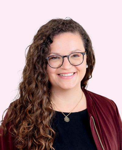 junge Frau mit lockigen braunen Haaren und runder Brille, burgunderfarbenem Blouson und filigraner Goldkette vor hellem rosa Hintergrund