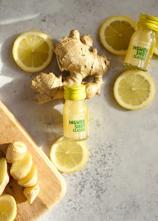 Zwei kleine Ingwershot-Fläschchen neben Ingwerknollen Zitronenscheiben auf hellem Marmoruntergrund