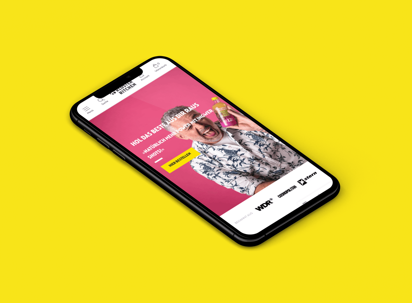 Schräg positioniertes Smartphone auf gelbem Hintergrund zeigt lächelnden Mann im Blumenhemd mit Ingwer-Shot auf rosa Hintergrund